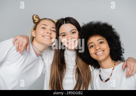 Porträt eines positiven jungen brünetten Teenagers, der zwischenrassistische Freundinnen in weißen T-Shirts umarmt und auf die Kamera schaut, isoliert auf grauem, energetischem teena Stockfoto