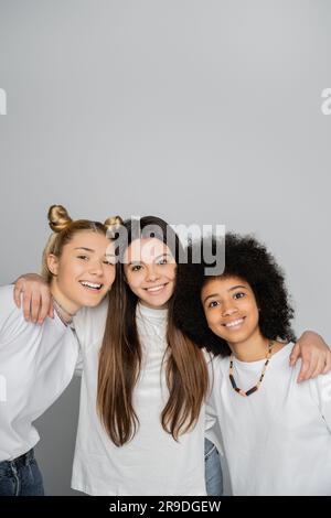 Lächelndes braunes Teenager-Mädchen in weißem T-Shirt, umarmt interrassistische Freundinnen und schaut zusammen in die Kamera, isoliert auf grauen, energiegeladenen Teenager-Models Stockfoto