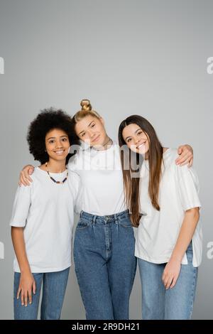 Stylische und fröhliche, gemischtrassige Teenager-Mädchen in weißen T-Shirts und blauen Jeans, die sich umarmen und in die Kamera schauen, während sie isoliert auf grauen Teenagern stehen Stockfoto