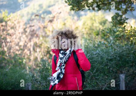 Eine afrikanische Frau in einem leuchtend roten Wintermantel steht vor dem Hintergrund üppiger grüner Vegetation Stockfoto