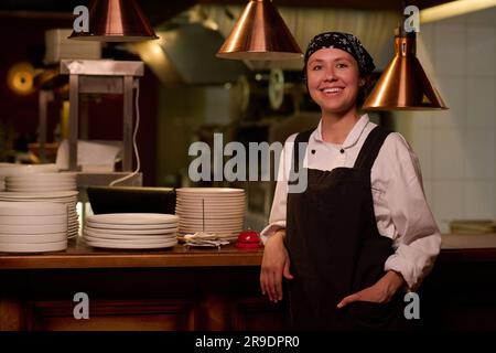 Junge, fröhliche Mitarbeiterin im Café oder Restaurant in Uniform, die vor die Kamera schaut, während sie mit Tellern an der Theke steht und auf Gäste wartet Stockfoto