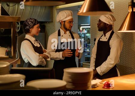 Drei junge interkulturelle Köche in Uniform diskutieren Arbeitspunkte in der Küche, während einer von ihnen Kollegen etwas erklärt Stockfoto