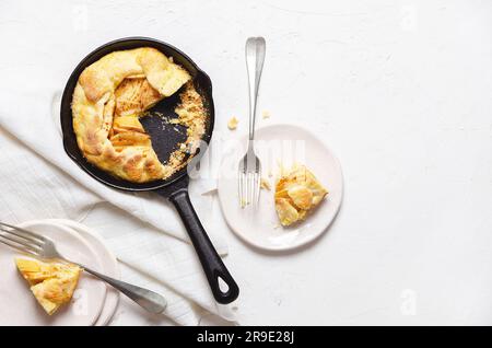 Apfelgalette in einer schwarzen Pfanne, zwei Galette in weißen Tellern mit Gabeln und ein Stück weißes Tuch auf weißem Hintergrund mit Kopierraum. Stockfoto