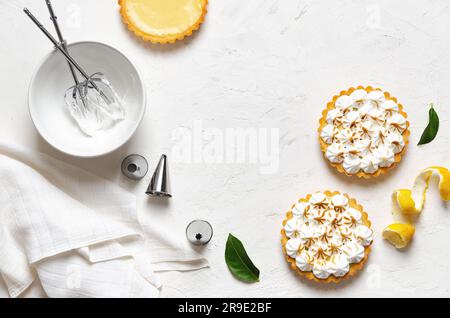 Mini-Zitronenkuchen mit Meringue, Zitronenschale, Zitronenblätter, Whisks in einer Schüssel mit Meringue, einigen Düsen und einer weißen Serviette auf weißem Hintergrund. Stockfoto