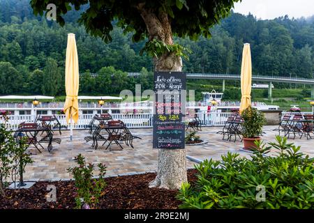 Eine Tafel an einem Baum wirbt für ein Gericht „nach altem DDR-Rezept“ und, wie es auf der Tafel heißt, „immer noch beliebt“ auf der Terrasse eines Hotels in Bad Schandau, Sachsen, Deutschland: „Zigeunersteak mit Pommes“ („Zigeunersteak mit Fritten“) – ein Begriff, der einst auf ostdeutschen Speisekarten üblich war und heute als diskriminierend gilt, da er von einem historischen Begriff für Sinti und Roma abgeleitet ist. Gelbe Sonnenschirme und leere Tische stehen auf der gepflasterten Terrasse, im Hintergrund spannt sich eine Brücke über die Elbe. Stockfoto