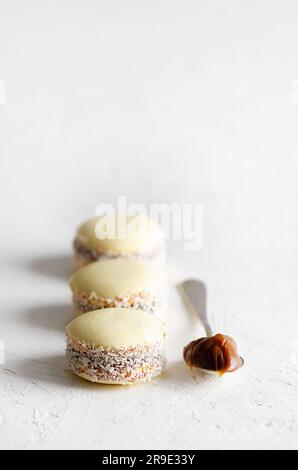 Drei Maisstärke-Alfajores mit Dulce de leche und Kokos und einem Löffel Dulce de leche auf weißem Hintergrund mit Kopierraum. Stockfoto