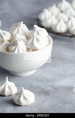 Weiße Meringue-Kekse in einer weißen Schüssel und auf einem schwarzen Gestell, auf grauem Hintergrund. Stockfoto