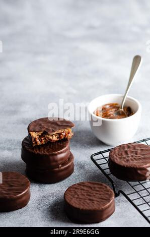 Dunkle Schokoladenalfajores auf einem dunklen Regal und eine Schüssel mit Dulce de leche auf grauem Hintergrund. Stockfoto