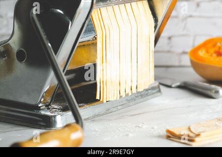 Butternusskürbis-Nudeln in einer Nudelmaschine, ein Messer und ein Stück Butternusskürbis, auf marmoriertem Hintergrund. Stockfoto