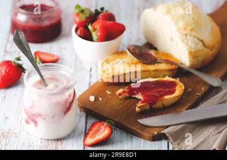 Ein Glas Joghurt, frische Erdbeeren, Erdbeermarmelade in einem Glas und auf einer Scheibe geröstetem Brot, auf hellem hölzernem Hintergrund. Stockfoto