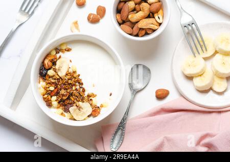 Joghurt mit Müsli, Banane, Nüssen und Rosinen und einer rosa Serviette auf einem weißen Tablett. Stockfoto