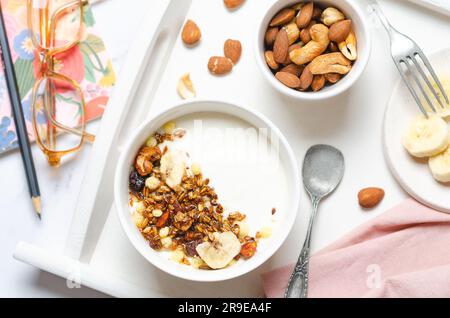 Gesundes Frühstück mit Joghurt, Müsli, Nüssen und Banane auf weißem Tisch. Stockfoto