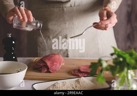 Eine Frau in einer beigefarbenen Schürze bringt Salz auf das Fleisch. Der Prozess der Herstellung argentinischer Milanesas. Stockfoto