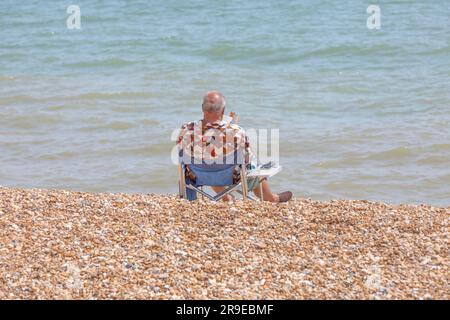 Ein Mann, der sich sonnt und ein Buch liest, sitzt auf einem blauen Liegestuhl an einem Kiesstrand. Stockfoto