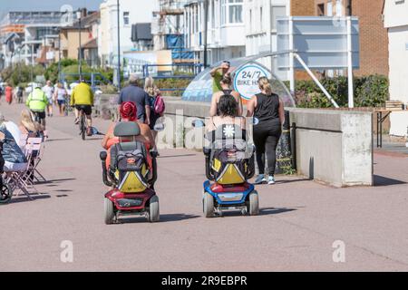 Zwei Damen auf Mobilitätsrollern, die entlang einer geschäftigen Strandpromenade fahren. Stockfoto