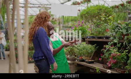 Seniorin, die jungen weiblichen Klienten in der Gartenwerkstatt hilft. Eine afroamerikanische ältere Frau, die Kunden beim Kauf von Pflanzen in einem kleinen Bus berät Stockfoto