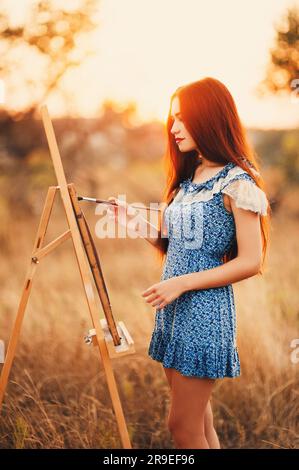 Junge, rothaarige Asiatin malt und kleidet auf einer Leinwand in der Natur Wiese bei Sonnenuntergang, elegante Frau in einem Naturhintergrund Stockfoto