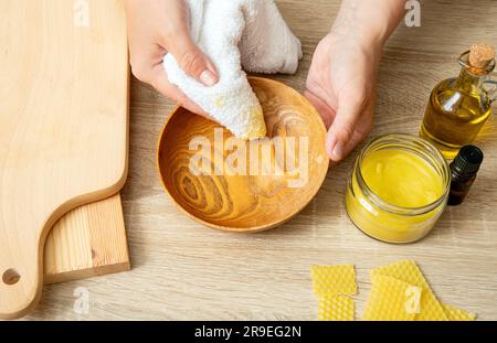 Frauenhände tragen hausgemachtes Bienenwachs-Holzpolitur auf, um die natürliche Farbe der Holzschüssel wiederherzustellen. Bienenwachs, Olivenöl und ätherisches Öl, weiches Tuch. Stockfoto