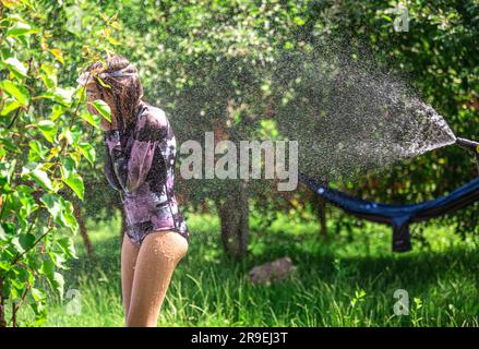 Lustiges kleines Mädchen, das im sonnigen Garten mit Gartenschlauch spielt. Stockfoto