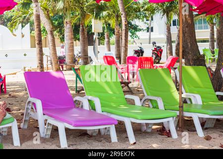 Schlammige, unordentliche Sonnenliegen am Strand von thailand warten auf Touristen. Sonnenliegen mit Matratzen in sanftgrünen und violetten Tönen unter Palmenschatten auf sandigen tropischen Plätzen Stockfoto