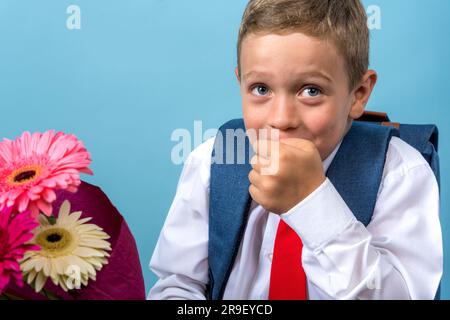 Ein glücklicher Erstklässler in einem weißen Hemd hält einen Blumenstrauß in den Händen, bedeckt seinen Mund verspielt mit seiner Hand und lacht. Ein süßes Kaukasien Stockfoto