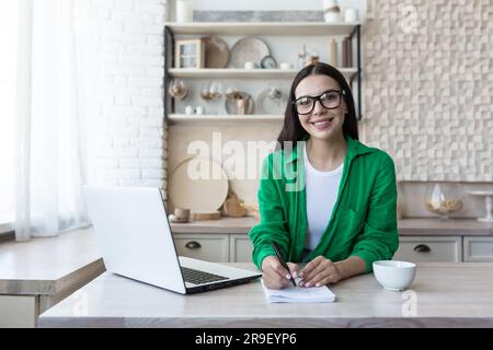 Porträt einer jungen Studentin, die per Fernstudium mit einem Laptop lernt. In der Küche am Tisch mit einem Notizbuch sitzen und lächelnd in die Kamera schauen. Stockfoto