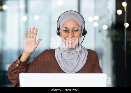 Nahaufnahme. Junge hispanische Studentin in Hijab, sitzt am Schreibtisch im Büro und trägt ein Headset und telefoniert per Videoanruf vom Laptop aus. Er winkt mit der Hand und sagt Hallo zur Kamera auf dem Monitor. Stockfoto