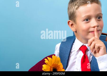 Ein glücklicher Erstklässler in einem weißen Hemd hält einen Blumenstrauß in den Händen und schließt spielerisch seinen Mund mit seinem Finger. Ein süßer weißer Junge Stockfoto