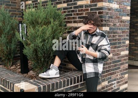 Ein junger Mann in der Stadt, der auf dem Bordstein sitzt und ein Telefon macht Stockfoto