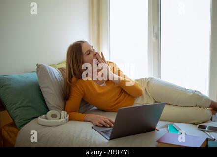 Die Verschlafene Dame Gähnt Sich Und Macht Hausaufgaben Auf Dem Laptop Im Schlafzimmer-Interieur Stockfoto