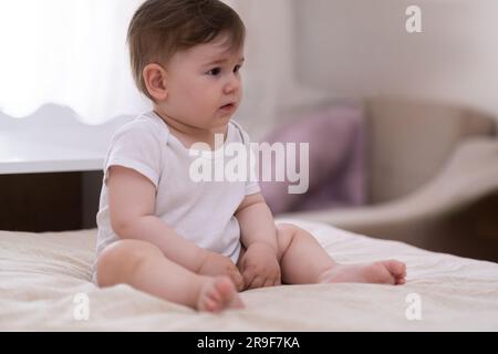 Süßer, dicker Junge im weißen Body, der auf einem weichen Bett sitzt und zur Seite schaut Stockfoto