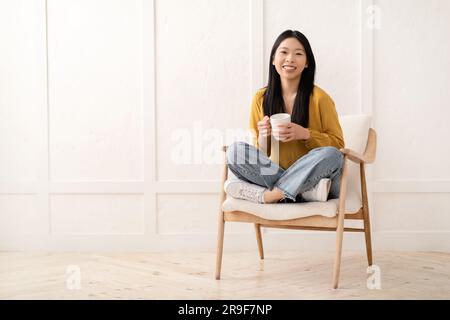 Fröhliche, hübsche asiatische Frau, die im Sessel sitzt und Kaffee trinkt Stockfoto