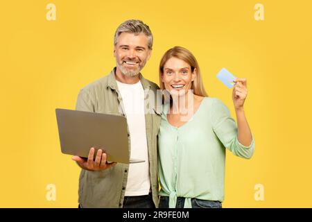 Glückliches Paar mittleren Alters in lässigen Outfits, Einkaufen und Banking im Internet, zeigt Bankkarte und benutzt Laptop Stockfoto