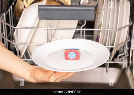 Weibliche Hand hält auf einem Teller ein Geschirrspültablett für eine Spülmaschine auf dem Hintergrund von schmutzigen Tellern, Geschirrspülern, Geschirrspülern Stockfoto