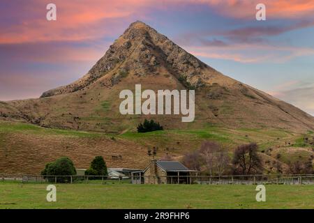 Foto einer alten kleinen Steinfarm in einem üppigen grünen Ackerfeld vor einem großen Hügel auf der Südinsel Neuseelands Stockfoto