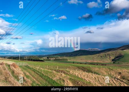 Foto eines großen Regenbogens über einem grünen landwirtschaftlichen Feld mit weidenden Kühen und Schafen und einer Bergkette im Hintergrund in Neuseeland Stockfoto