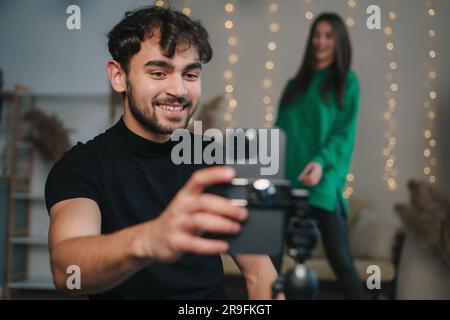 Fröhlicher junger bärtiger Mann in lässiger Kleidung, der Selfie mit dem Handy macht, während er in einem gemütlichen Zimmer am Tisch sitzt Stockfoto