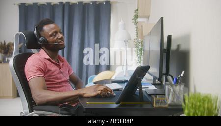 Afroamerikanischer Webdesigner oder -Entwickler arbeitet Remote zu Hause. Der Mann sitzt und spricht auf einer Online-Konferenz am Computer. Tablet und mehrere Monitore stehen auf dem Tisch. Das Konzept der Arbeit im gemütlichen Zuhause. Stockfoto