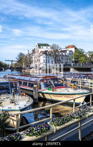 Tourboote, die an der Amstel im Rembrandt Plein in Amsterdam anlegen, mit Aluminiumbrücke im Hintergrund Stockfoto