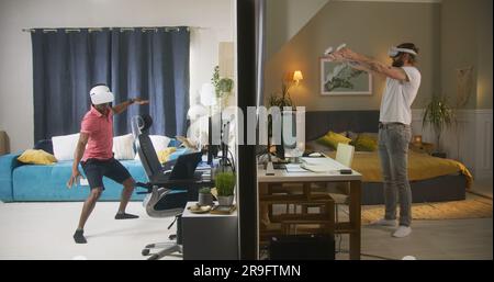 Ethnische Männer und Nachbarn in VR-Headsets spielen Augmented-Reality-Videospiele mit Wireless-Controllern. Blick auf zwei durch Wände getrennte Zimmer oder Apartments. Konzept von Nachbarschaft und Lebensstil. Stockfoto