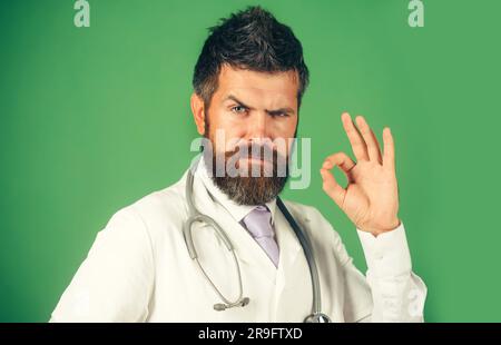 Ein ernster Arzt zeigt ein gutes Zeichen. Bärtiger Mann mit Stethoskop im weißen Kittel mit Zeichen-in-Zulassung. Medizin, Klinik, Behandlung, Gesundheitswesen Stockfoto
