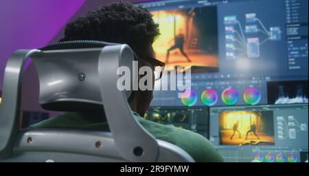 Rückansicht eines afroamerikanischen Redakteurs, der Farbkorrektur macht, im Heimstudio arbeitet. Actionfilmaufnahmen und Software-Schnittstelle auf Computer und großem digitalen Bildschirm. Das Konzept der Filmpostproduktion. Stockfoto