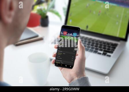 Der Kerl konzentrierte sich auf die Wettquoten auf seinem Smartphone. Folgt einem Fußballspiel auf seinem Laptop Stockfoto