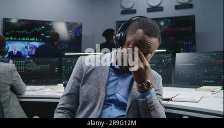 Ein müder afroamerikanischer Finanzanalyst, der am Arbeitsplatz sitzt, nimmt die Brille ab und schaut in die Kamera. Kollegen analysieren Börsenkurse auf Bildschirmen im Hintergrund. Investitionskonzept. Porträt. Stockfoto