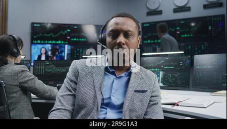 Ein müder afroamerikanischer Finanzanalyst, der am Arbeitsplatz sitzt, nimmt die Brille ab und schaut in die Kamera. Kollegen analysieren Börsenkurse auf Bildschirmen im Hintergrund. Investitionskonzept. Porträt. Stockfoto