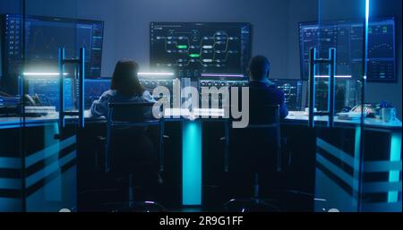 Rückblick auf verschiedene IT-Technikspezialisten, die nachts an Computern mit Datenbankserver in modernen Überwachungsbüros arbeiten. Große digitale Bildschirme mit Echtzeit-Analysediagrammen und Blockchain-Netzwerk. Stockfoto