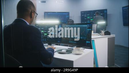 Männlicher afroamerikanischer Ingenieur tippt Tastatur und analysiert angezeigte Serverdaten und Echtzeitdiagramme. Multikulturelle Big-Data-Spezialisten arbeiten im Hintergrund. Konzept der Cybersicherheit. Rückansicht. Stockfoto