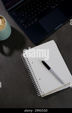 Draufsicht auf den Arbeitsplatz mit schwarzem Laptop, leerem Notebook und grüner Tasse Kaffee Stockfoto