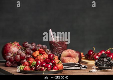 Stillleben mit roten Pflaumen, paraguayern, Trauben, Waldfrüchten, Kirschkirschen und Entgiftungssaft auf einer alten Metallplatte und einem Hintergrund aus Holz-pla Stockfoto