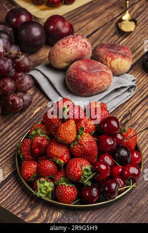 Stillleben mit roten Pflaumen, paraguayern, Trauben, Beeren, reifen Kirschen, Auf einer alten Metallplatte und einem Hintergrund aus dunklen Holzplanken Stockfoto